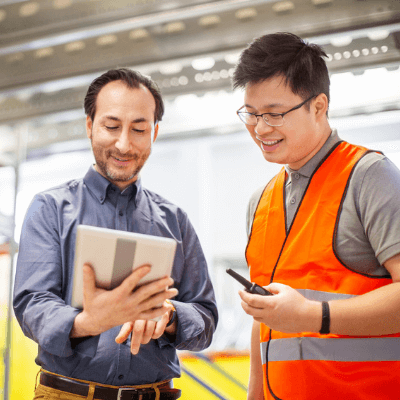 Two warehouse workers talking together over a tablet while standing inside of a large warehouse